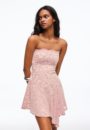 Kobieta w różowej koronkowej sukience bez ramiączek z kwiatowym wzorem, patrząca w lewo na jednolitym białym tle.