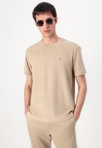 Beige lyhythihainen t-paita pehmeästä kankaasta pyöreällä kauluksella, jossa on pieni brodeerattu logo rinnassa. Yhdistetty vaaleanbeigeihin housuihin.