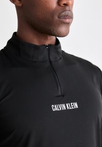 Nærbilde av en person som har på seg en svart kvart-zip topp med "CALVIN KLEIN" logo plassert på brystet.