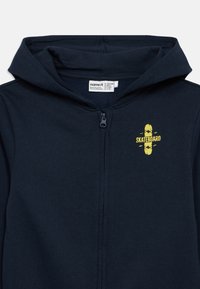 Tmavomodrá mikina s kapucňou a predným zipsom, s grafikou skateboardu v žltej farbe. Vyrobená z mäkkého materiálu, s kapucňou pre teplo.