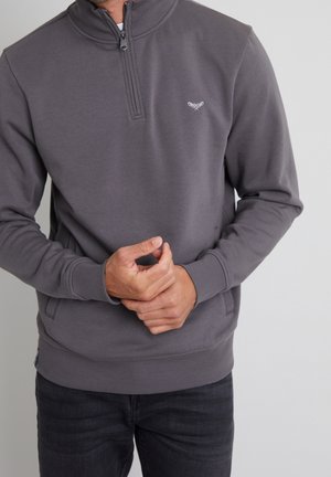 Mann trägt einen dunkelgrauen Sweatshirt mit Viertel-Reißverschluss und kleinem weißem Sticklogo auf der Brust, die Hände vor dem Körper verschränkt.