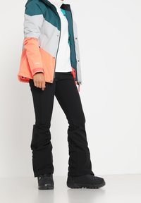 Chaqueta aislante colorida con secciones en teal, gris y naranja. Llevada sobre un suéter blanco, combinada con pantalones de esquí negros y botas de invierno negras.