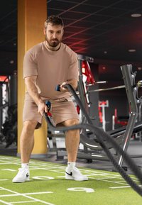 Completo sportivo beige con maniche corte e pantaloni corti, che afferra corde da battaglia nere su una superficie di allenamento verde in una palestra con attrezzature per esercizi.