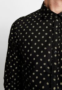 Camicetta nera con bottoni, caratterizzata da un motivo a piccole foglie in verde chiaro ripetuto. Il tessuto sembra liscio e presenta un colletto standard.