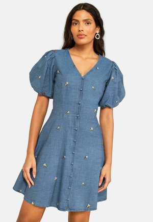 Naine kannab sinist krepste varrukatega kleiti, millel on väikesed tikitud lilled ja kangaga kaetud nööbid ees.