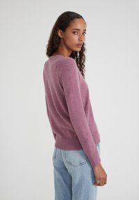 Eine Frau mit lockigem Haar trägt einen mauvefarbenen Pulli und hellblaue Jeans und steht seitlich vor einem schlichten weißen Hintergrund.