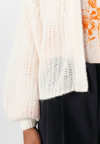 Cardigan tricotat în crem, cu un design lejer și aerisit, având mâneci bufante și manșete cu model în dantelă, purtat deasupra unei bluze cu model portocalie.