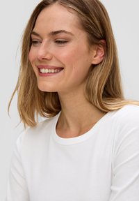 Weißes, geripptes Langarmshirt mit Rundhalsausschnitt. Die Textur ist glatt und der Stoff scheint leicht zu sein. Das Modell lächelt.