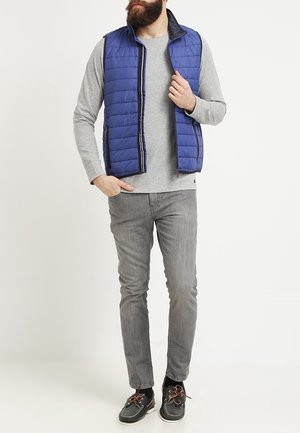 Mężczyzna w niebieskiej pikowanej kamizelce na szarej, długorękawowej koszuli, szarych dżinsach i czarnych butach typu boat stoi na białym tle.