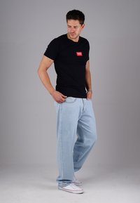 Man standing wearing black t-shirt with red logo, light blue jeans, white sneakers, looking down and to the side against plain gray background.