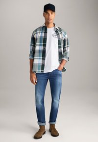 Karoviertes Hemd in Grün und Weiß, weißes T-Shirt, blaue Jeans, braune Stiefel. Lässiges Outfit mit Knöpfen und hochgekrempelten Ärmeln am Hemd.