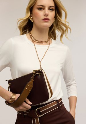 Blonde Frau trägt ein weißes Oberteil und braune Hosen mit einem gestreiften Gürtel, hält eine braune Leder-Clutch und trägt goldenen Schmuck.