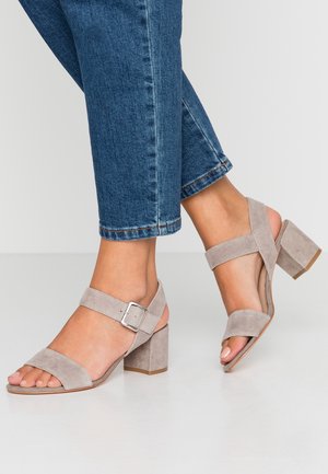 Pieds chaussés de sandales à talons blocs en daim beige avec des brides à la cheville et des boucles argentées, associées à un jean en denim bleu raccourci.