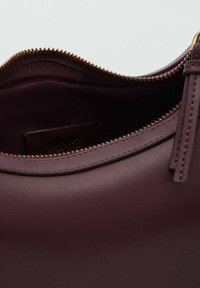 Sac en cuir bordeaux à texture lisse, doté d'une fermeture éclair dorée et d'une languette de tirage. L'intérieur révèle une doublure en tissu avec une étiquette de marque.
