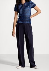 Persona con una camisa polo de manga corta a rayas azules, pantalones oscuros de corte ancho y planos de ballet blancos, de pie con una mano en la cadera.