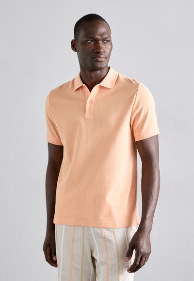 Tricou polo piersici cu guler clasic, mâneci scurte și un logo subtil pe piept, asociat cu pantaloni cu dungi de culoare deschisă.