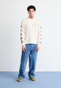 Camisa de manga longa canelada em bege claro com gráficos pretos nas mangas. Combinada com calças de ganga largas em azul e tênis verdes.