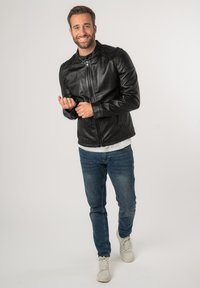 Schwarze Lederjacke mit Reißverschluss, Stehkragen und Seitentaschen. Getragen über einem weißen Hemd und blauen Jeans mit Turnschuhen.
