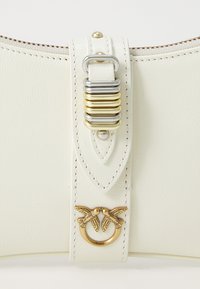 Borsa in pelle bianca con una tracolla centrale dotata di accessori in oro e argento. Dettagli notevoli includono un emblema a doppio uccello e cuciture a contrasto.