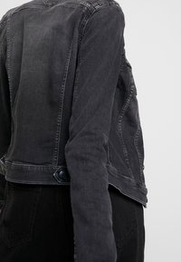 Czarna kurtka dżinsowa o dopasowanym kroju, z widocznymi szwami, guzikami jako akcentami oraz lekko wyblakłym wykończeniem, podkreślającym casualowy styl.