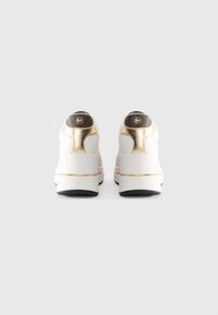 Pár bielych platformových tenisiek so zlatými akcentmi a tmavohnedými logovými nášivkami na päte, zobrazené zozadu na šedom pozadí.