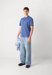 Zila kokvilnas T-kreklu ar krūšu kabatu, apvienots ar gaiši ziliem platām džinsiem un melniem apaviem. Krekliņš satur logotipa detalizāciju uz kabatas.