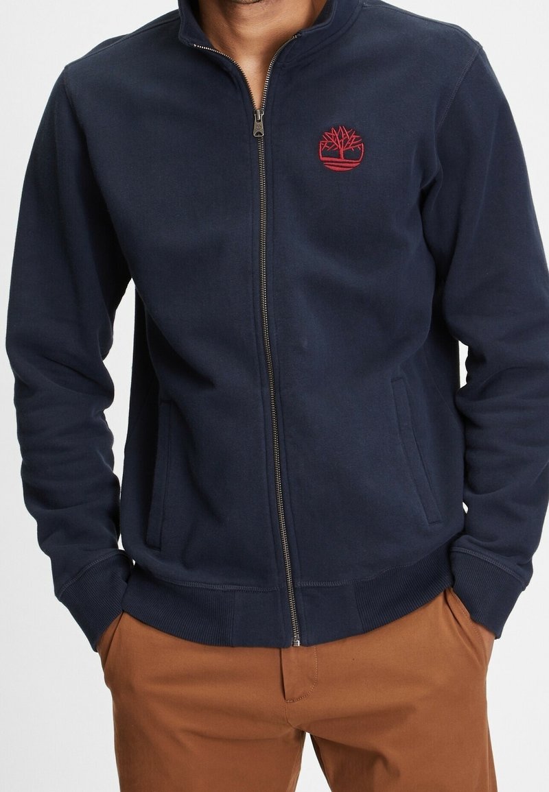 Homme portant une veste zippée bleu marine avec un logo d'arbre rouge sur la poitrine et un pantalon marron, les mains dans les poches, sur fond blanc.