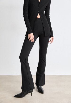 Cárdigan negro de manga larga con botones y una abertura frontal; combinado con pantalones de campana negros ajustados y botas negras de tacón puntiagudo.