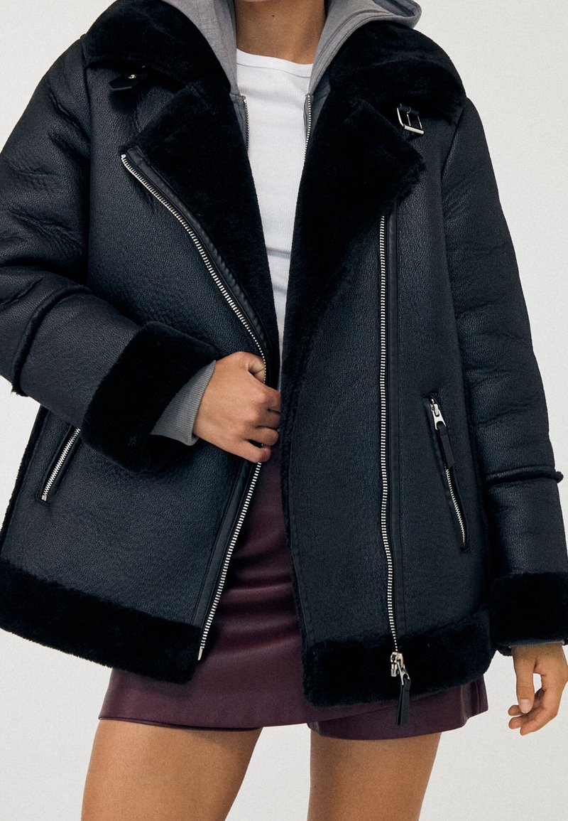Personne portant une veste en peau lainée noire avec des fermetures éclair et une boucle sur un haut blanc et une jupe en cuir rouge foncé.