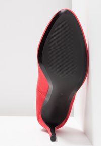 Chaussure à talon haut en daim rouge avec un bout pointu, dotée d'une semelle en caoutchouc noir lisse et d'un dessous texturé pour l'adhérence.