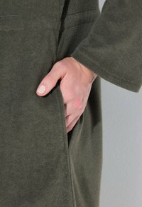 Zelená texturovaná látka s detailem kapsy. Ruka je umístěna uvnitř kapsy, na níž je vidět světlý lak na nehty.