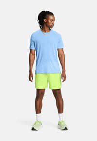 Camiseta deportiva de color azul claro de manga corta, combinada con pantalones cortos de un brillante color verde neón. El modelo lleva calcetines blancos y zapatillas de correr de colores claros.