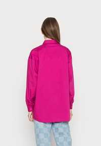 Kvinna med långt brunt hår står vänd bort, iklädd en ljusrosa långärmad tröja och ljusblå rutiga byxor mot en enhetlig bakgrund.