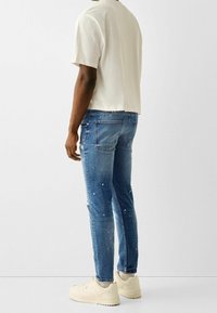 Svetle béžové tričko s kulatým výstřihem, modré obnošené džíny s barvou na skvrnách a krémové tenisky. Viditelné švy a kapsy na džínách.