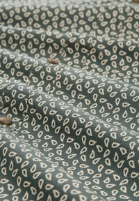 Tissu vert avec un petit motif de feuilles blanches, présentant des coutures et des boutons visibles, surface texturée avec un drapé doux.