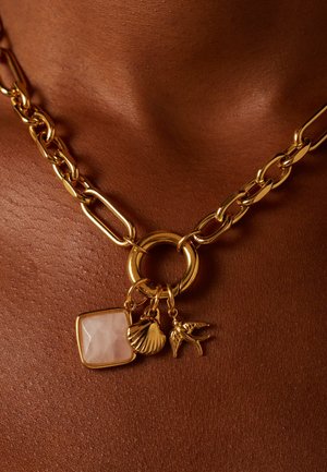 Collier en chaîne dorée avec de gros maillons, un anneau central, et trois pendentifs : une pierre carrée, une coquille et un oiseau, sur un fond de teint.