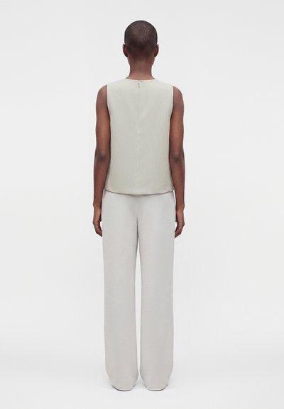 Personne debout de dos, portant un haut sans manches beige clair et un pantalon assorti à jambes larges, sur fond blanc uni.