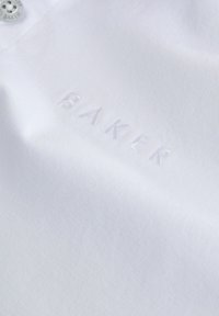 Bílá bavlněná látka s jemnou texturou, na které je embosované slovo "BAKER" v světlé barvě, doplněné stříbrným knoflíkem.