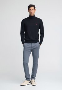 Maglione nero con collo alto e polsini a coste, pantaloni aderenti grigi e sneaker beige. Piccolo dettaglio del logo rosso sul petto.