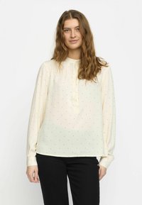Blusa crema con maniche lunghe, patta abbottonata e piccole pois neri. Tessuto morbido con una vestibilità ampia e orlo arrotondato. Abbinata a pantaloni neri.