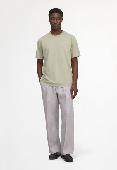 Homme debout portant une chemise à manches courtes vert clair, un pantalon plissé gris clair et des mocassins noirs sur fond blanc.