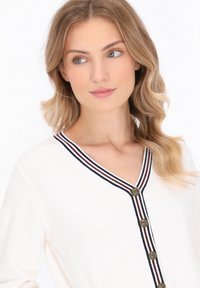 Vrouw met golvend blond haar in een witte V-hals sweater met marineblauwe en rode gestreepte afwerking en gouden knopen, kijkt naar de zijkant.