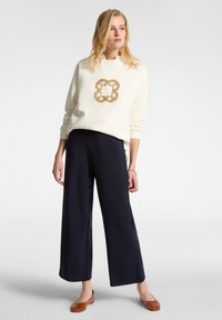 Krémová mikina s zlatým květinovým vzorem, doplněná o tmavě modré kalhoty s širokým střihem a hnědé ploché boty. Hladká tkanina, volný střih po celé ploše.