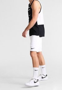 Canottiera atletica bianca e nera, pantaloni corti bianchi, calze bianche con dettagli neri e scarpe da ginnastica bianche con logo nero.