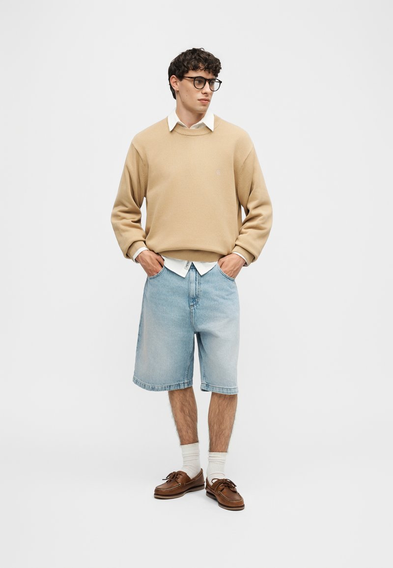 Jeune homme avec des lunettes portant un pull beige sur une chemise blanche, un short en jean clair jusqu'aux genoux, des chaussettes blanches et des mocassins marron, debout les mains dans les poches.