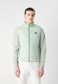 Chaleco acolchado de color verde claro con cuello alto, bolsillos con cremallera y un parche de logo. Lleva puesto sobre una camiseta de manga larga de color gris claro.