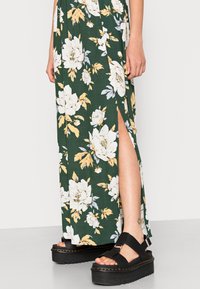 Robe maxi florale avec un fond vert, des fleurs blanches et jaunes, et une fente haute sur la cuisse. Sandales à plateforme noires avec un détail de sangle.