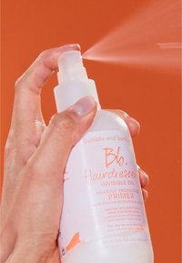 En klar sprayflaska med en svart pumpmunstycke. Innehåller 250 ml hårprimer märkt "Invisible Oil" i orange text på vit bakgrund.