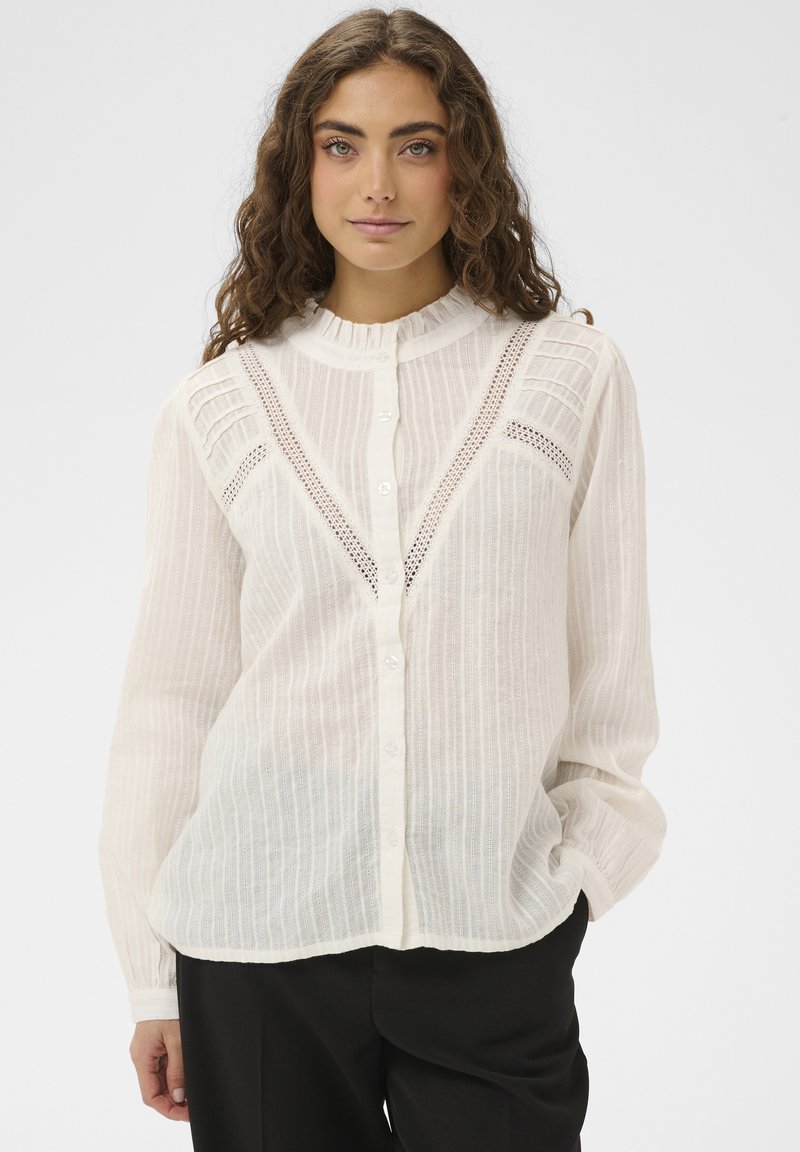 Blouse blanche légère à boutons avec des rayures verticales, un col à volants et des accents en dentelle sur la poitrine ; manches longues avec poignets froncés.
