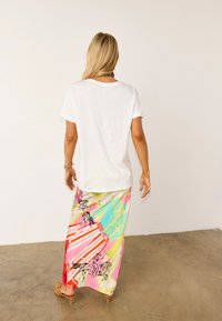 Γυναίκα με μακριά ξανθά μαλλιά, φορώντας λευκό μπλουζάκι και πολύχρωμη, εμπριμέ μακριά φούστα, στέκεται σε τσιμεντένιο δάπεδο μπροστά σε λευκό απλό τοίχο.